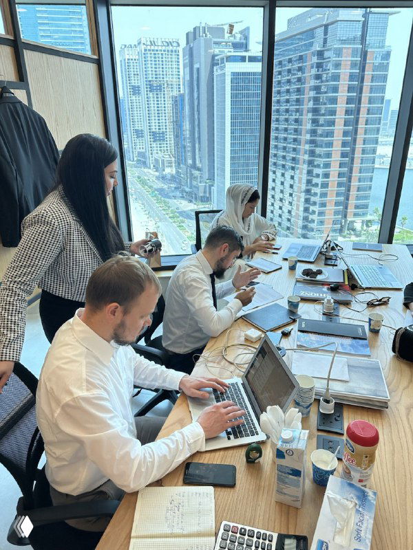 Команда маркетологов обсуждает работу за столом с ноутбуками и заметками, большое окно с видом на высотки города на заднем плане.