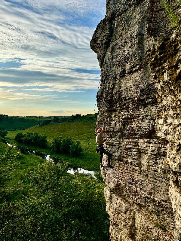 Скалолаз на отвесной скале, вид сбоку: спортсмен взбирается над зелёной долиной и рекой, подчёркивая масштаб и высоту подъёма.
