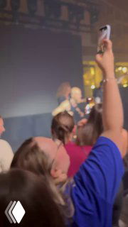 Зал конференции: аудитория снимает сцену на смартфоны — размытый кадр с поднятыми руками и выступающим на сцене.