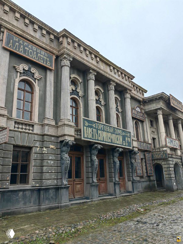 Фасад большого здания с колоннами и вывеской в декорациях, реплика городской архитектуры для съёмок на территории Мосфильма под пасмурным небом.