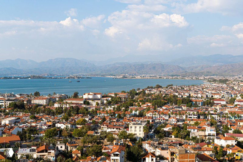 Широкая панорама города Фетхие с высоты: плотная прибрежная застройка с красными крышами, марина у залива и горы на дальнем плане под переменчивым небом.