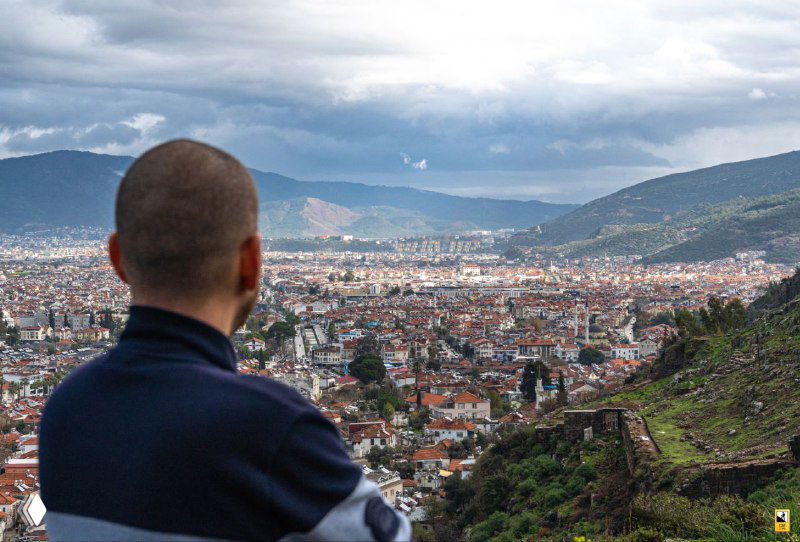 Человек сзади на смотровой площадке, смотрит на город Фетхие и залив; в кадре видна крепость на склоне и протянувшаяся городская застройка.