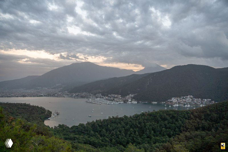 Вид на марину и бухту с высоты: плотная зелёная растительность на берегах, стоянка яхт в заливе и спокойная морская гладь в вечернем свете.