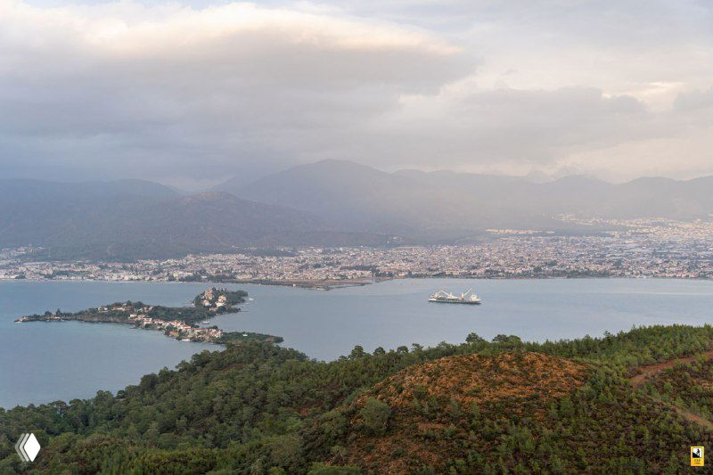 Панорамный вид на залив с островом и набережной: белые суда в бухте, городская линия вдоль берега и горы на заднем плане создают типичную прибрежную сцену.