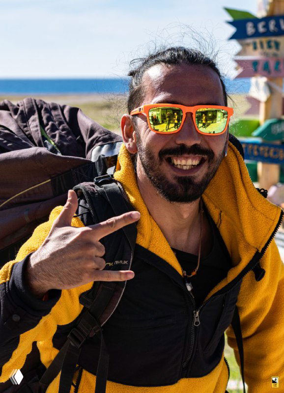 Улыбающийся инструктор по параглайдингу в жёлтой куртке и солнцезащитных очках на фоне моря и пляжного пейзажа с рюкзаком на плечах.