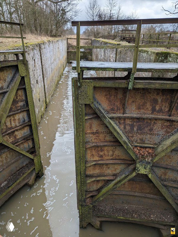 Вид сверху на железные перекладины и узкий водный коридор шлюза №2; мутная вода, мох и облупившаяся краска на металле.