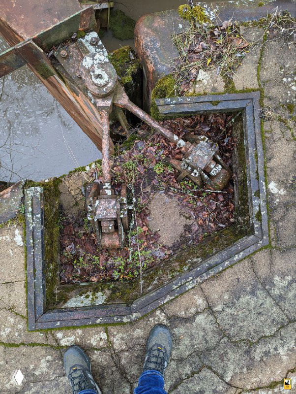 Механизм управления шлюзом с корродированными деталями, внутри — листья и мусор; внизу видны ботинки автора для масштаба.