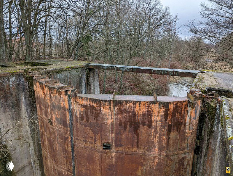 Ржавый изогнутый торец створки шлюза, выраженная коррозия и зелёный налёт мха на поверхности металла и бетона на краю лотка.