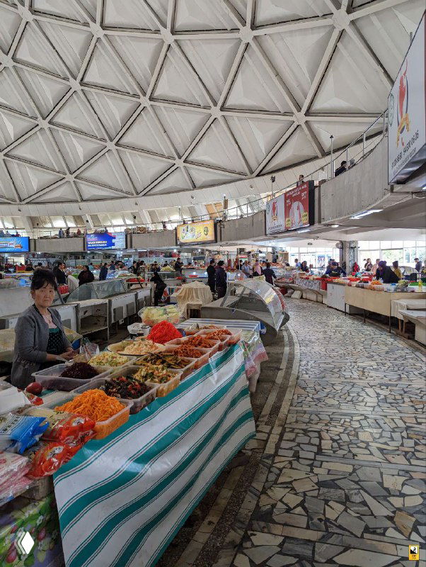 Круглая крытая часть рынка с торговыми прилавками внутри: соленья, сухофрукты и продукты аккуратно выложены, торговцы за прилавками и мозаичный пол заметны в кадре.