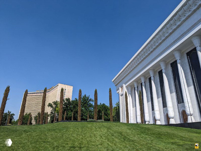 Белая колонная постройка и зелёный газон в городской среде Ташкента; сочетание классических форм и советской эпохи на фоне современного города.