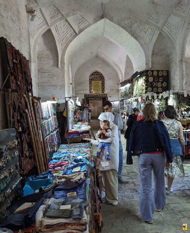 Внутри крытого базара: торговцы, прилавки с тканями и сувенирами, туристы и местные жители в оживлённой коммерческой атмосфере.
