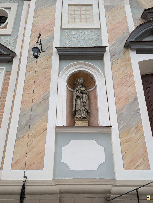 Фрагмент фасада костела с нишей и статуей святого, выполненной в барочном стиле, на фоне пастельной окраски стен и лепных деталей.