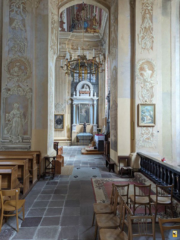 Боковой проход церкви с деревянными скамьями и лепниной на стенах, вид на интерьерный коридор и элементы художественного убранства.