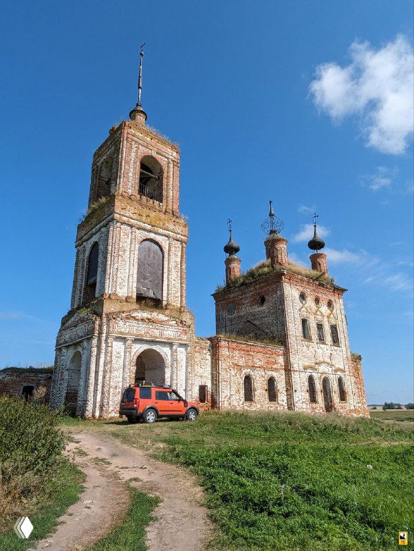 Разрушенная кирпичная церковь Флора и Лавра в селе Кибол на фоне неба; у входа виден припаркованный красный автомобиль, окружение полей.