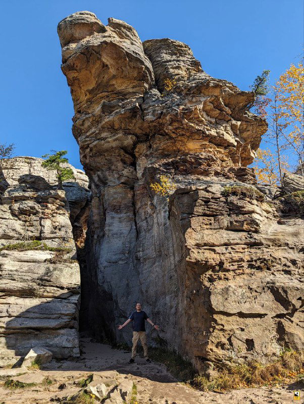 Песчаниковая вертикальная скальная формация с узким арочным проходом в основании и человеком для масштаба на фоне голубого неба.