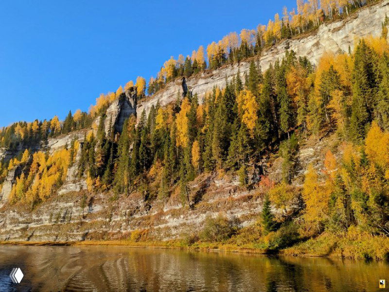 Осенняя панорама реки Усьва с высокой каменной стеной, желто-зелёными деревьями на склоне и зеркальным отражением в спокойной воде.