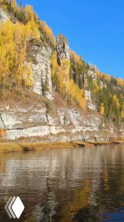 Крупный фрагмент скалы и берега Усьвы в осенней окраске; вид сбоку на каменные образования, листва и спокойную воду под голубым небом.