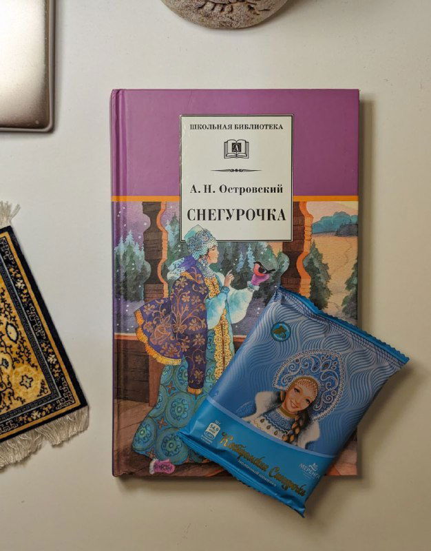 Обложка пьесы Александра Островского «Снегурочка» на столе рядом с шоколадкой с изображением девочки и тканым сувениром в русском стиле.