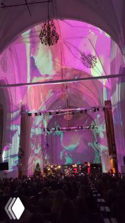 Интерьер Кафедрального собора с разноцветными проекциями и сценой; видно публику и музыкальную группу, создающую рок-версию классических тем.