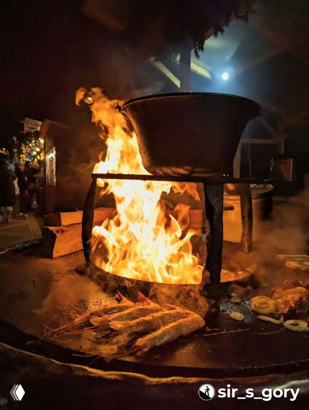 Огромный котёл на открытом огне на уличном новогоднем рынке: жар, дым и процесс приготовления морепродуктов и блюд на фоне праздничных палаток.