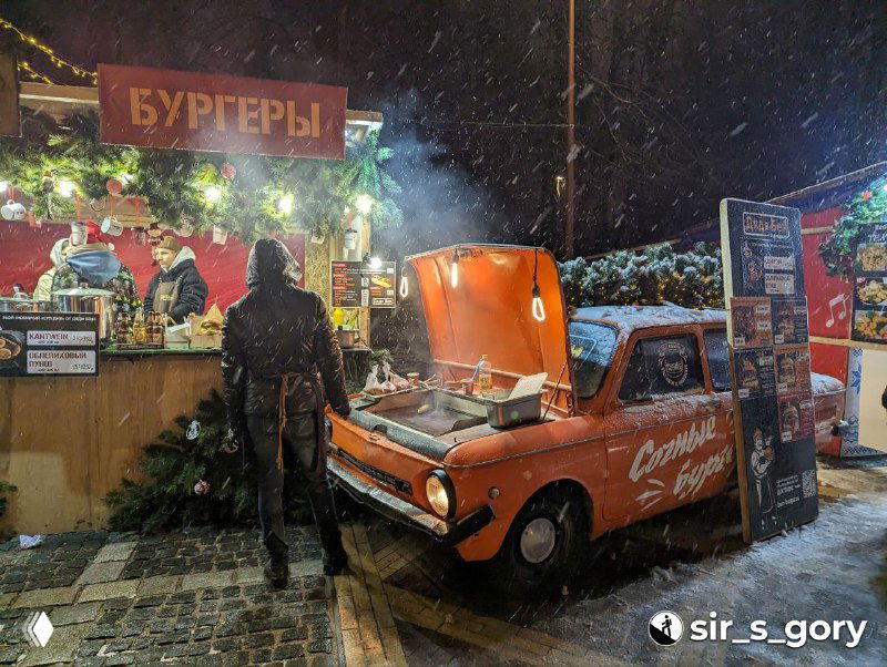 Фургон с открытым капотом, рядом палатка с бургерами на капоте и праздничным декором — сцена уличной еды на острове Канта в новогоднюю ночь.