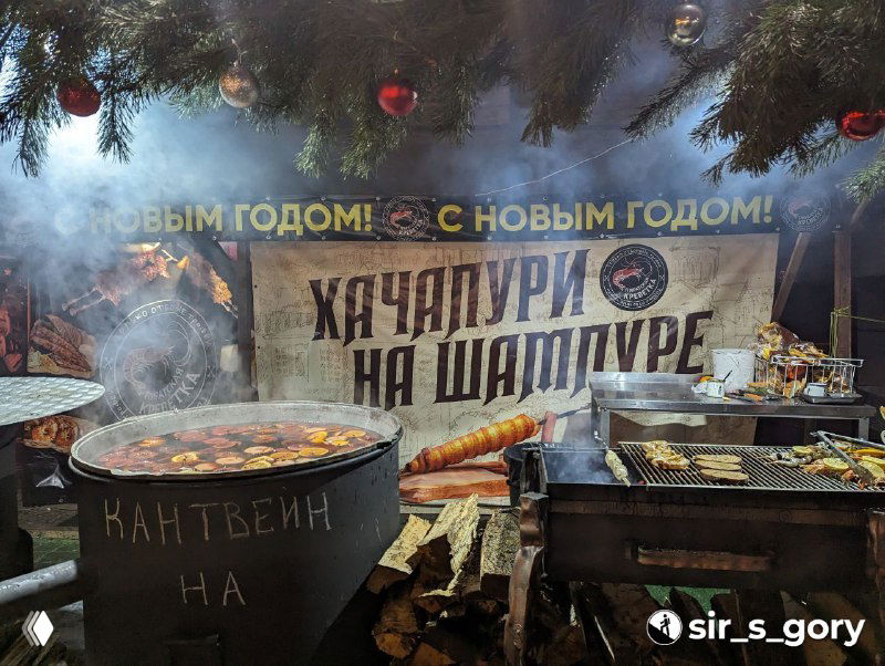 Крупная чаша с надписью "кантвейн" и открытым огнём, рядом гриль и хачапури на шампуре на фоне праздничного стэнда с вывеской и огнями.