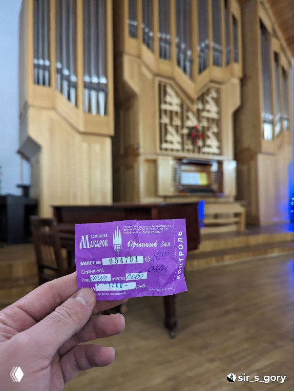 Билет на концерт в органном зале на переднем плане, в глубине виден интерьер зала и трубы органа — кадровый ракурс с рукой держателя билета.