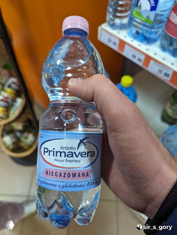 Рука держит бутылку польской воды Primavera 1 л в магазине: выпуклая этикетка на пластиковой таре и полки с товарами на фоне.