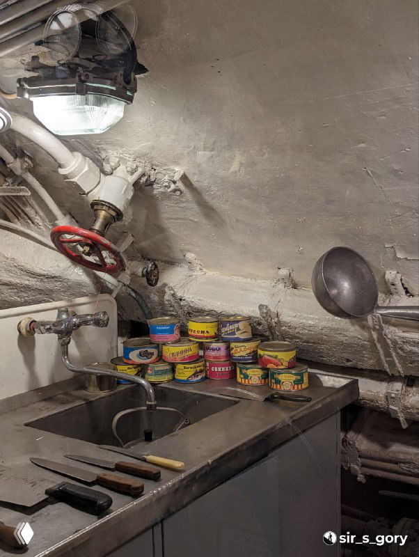 Камбуз подводной лодки: мойка, стопка консервных банок и кухонные принадлежности в тесном помещении, иллюстрирующие быт экипажа на борту.