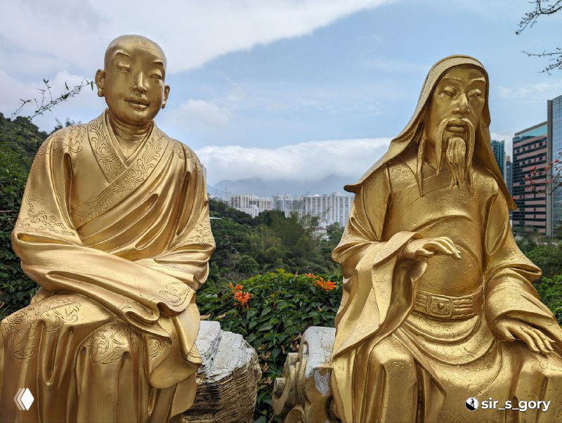 Две позолоченные статуи Будд на переднем плане с видом на зелёный склон и урбанистический пейзаж Гонконга позади, спокойная сцена.