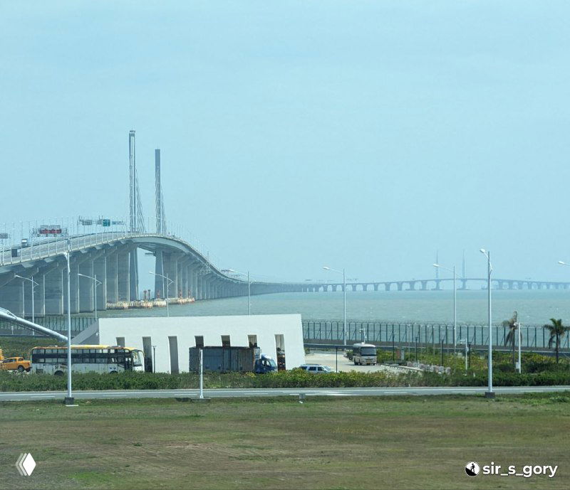 Панорамный вид на морской мост Гонконг—Чжухай—Макао с береговой точки: дорожная полоса, опоры моста и спокойное море под светлым небом.