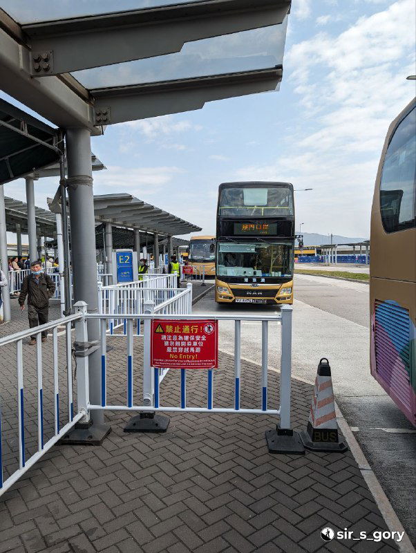 Автобусный терминал у въезда в Макао: двухэтажный автобус подъезжает к посадочной зоне, ограждения, указатели и ожидающие пассажиры на платформе.