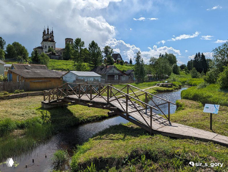 Деревянный пешеходный мост и настил над небольшой рекой в Тотьме на фоне старинной церкви и зелёных холмов в солнечный летний день.