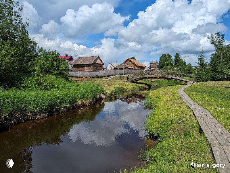 Речная заводь с отражением неба и деревянной тропой вдоль берега, рядом видны частные дома и ограждения традиционной деревянной застройки Тотьмы.