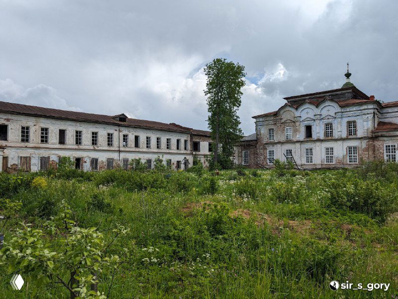 Комплекс монастырских зданий и корпуса с заросшей травой на переднем плане, кирпичные стены, окна и остатки хозяйственных построек на территории.