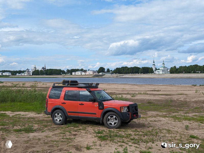 Красный внедорожник на фоне широкого песчаного берега и набережной Великого Устюга, видна противоположная часть города и церковные силуэты под небом.