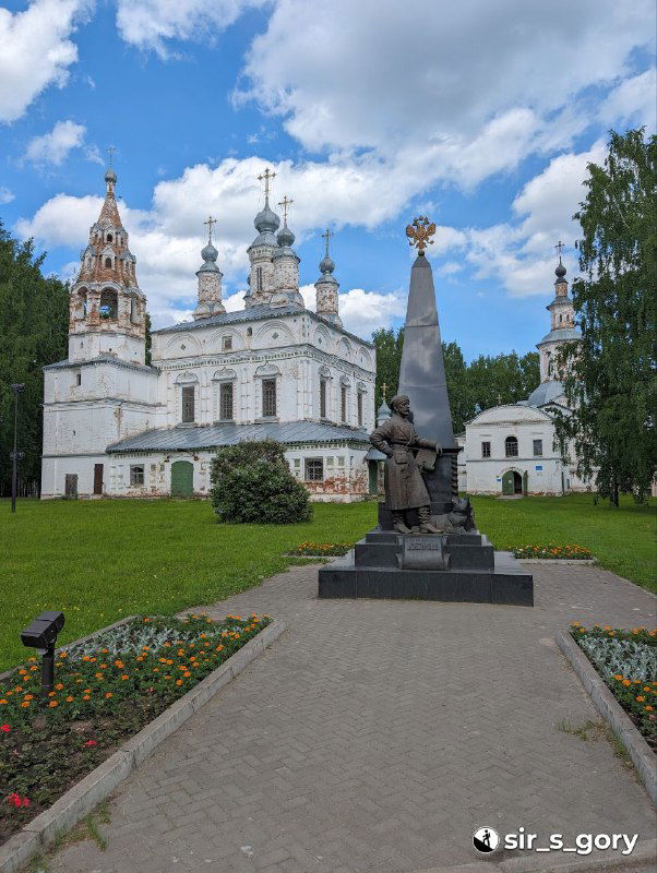 Памятник и сквер на подходе к Михайло-Архангельскому монастырю, белые стены и башни монастыря на фоне зелёной лужайки и цветников летом.