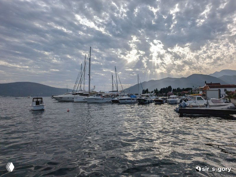 Портовая панорама с яхтами и моторными лодками на воде при облачном небе; в кадре видна береговая линия курорта и серебристые блики на море.