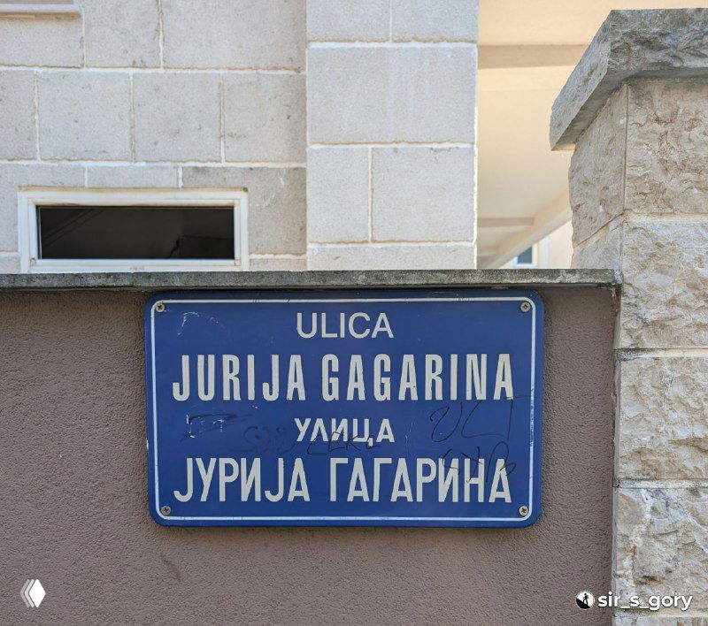 Уличная синяя табличка «Ulica Jurija Gagarina» с надписью латиницей и кириллицей на каменной стене — пример локальных топонимов в Черногории.