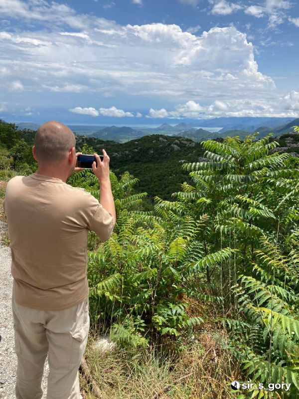Путешественник стоит спиной и фотографирует зелёные холмы и дальние горы: видовой кадр с наблюдением за ландшафтом Черногории и туристической активностью.