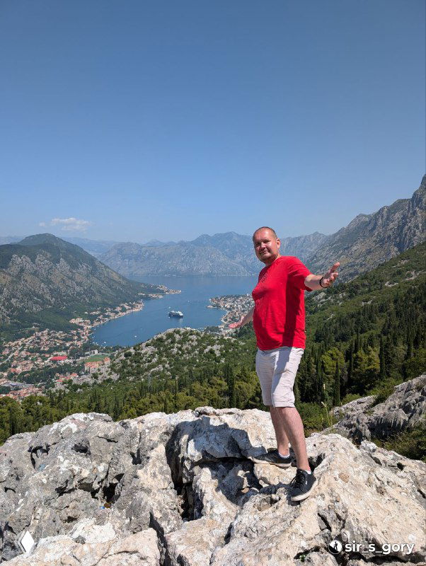 Турист в красной футболке стоит на каменной осыпи на фоне Которского залива — фигурный план на фоне панорамного вида.