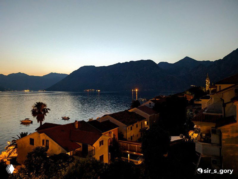 Вечерний вид на набережную и горы в сумерках: огни, силуэты крыш и спокойная вода залива в тёплых тонах заката.