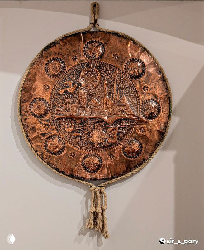 Круглый медный предмет с чеканным орнаментом и сценой в центре — либо щит, либо поднос, представленный как образец прикладного искусства в музее.
