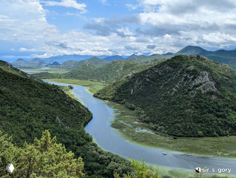 Панорама Скадарского озера: извилистая лагуна между зелёными холмами и Динарским нагорьем, мягкий свет и облака в небе.