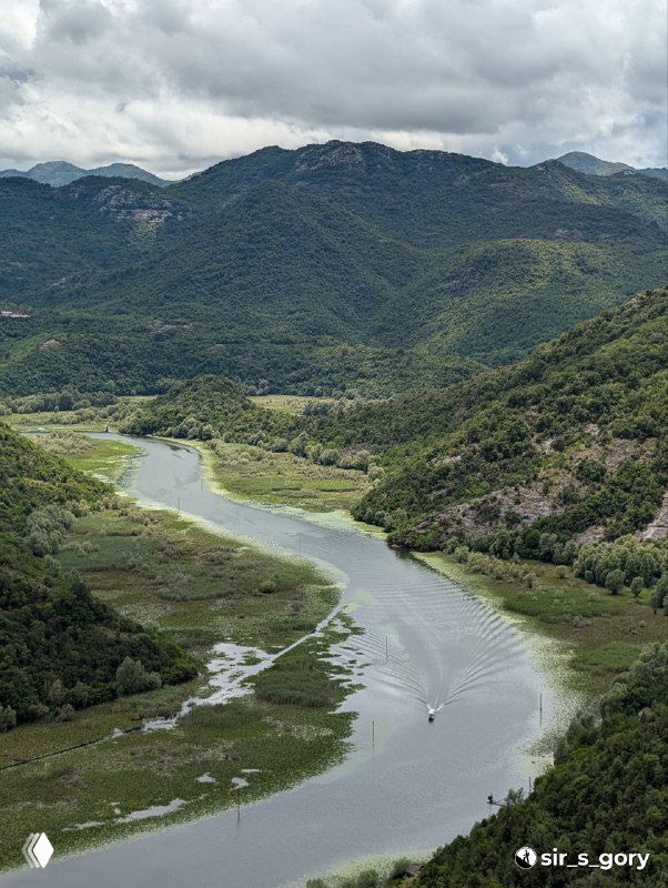 Река, втекающая в озеро, вид сверху с зелёных склонов; лодочная дорожка на воде и мелководные участки между островками.