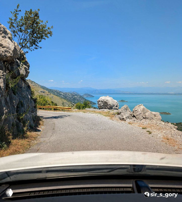 Узкая горная дорога у обрыва с видом на бирюзовое озеро; в кадре фрагмент капота автомобиля и скалистые откосы.