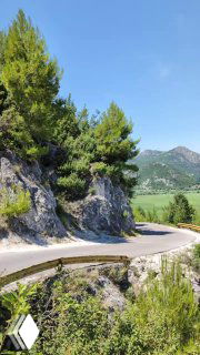 Извилистая серпантинная дорога у сосновых скал с видом на озёрные луга и холмы; летняя зелень и чистое синее небо.