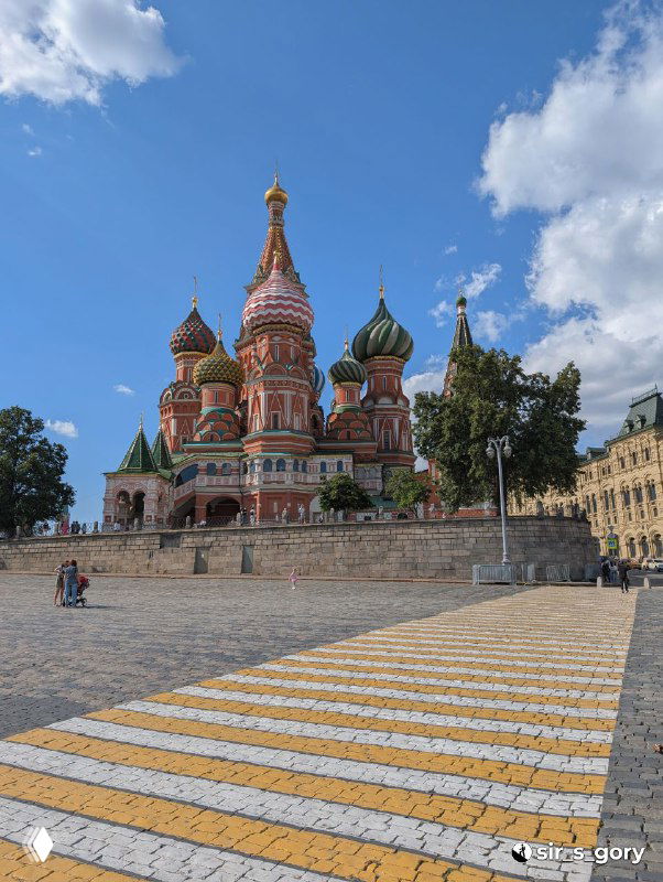 Панорамный вид Покровского собора на Красной площади: разноцветные луковичные купола, голубое небо и туристы на площадке перед храмом.