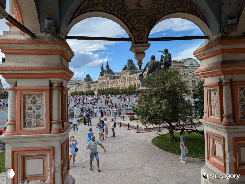 Вид через арку на Красную площадь: люди на площади, фасады зданий и часть убранства входа в Покровский собор, создающие городской контекст.