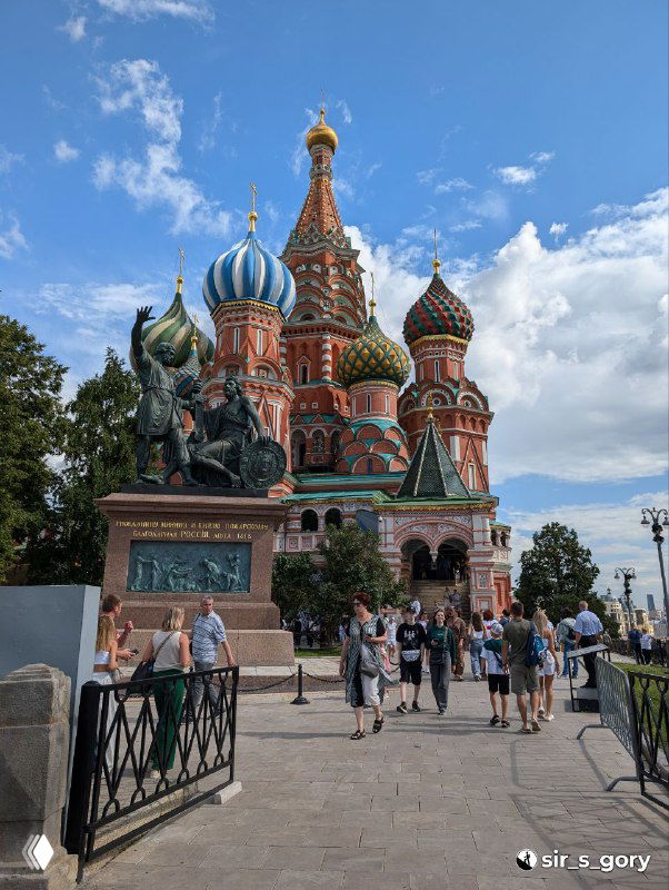 Общий кадр Покровского собора с памятником Минину и Пожарскому на переднем плане, туристы у ограждения и разноцветные купола на фоне облаков.
